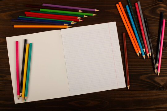 School notebook with oblique lines, colored pencils on the table wooden background. Blank sheet of paper in notebook on the desk. Back to school. School supplies. Top view. Copy space for text - Powered by Adobe