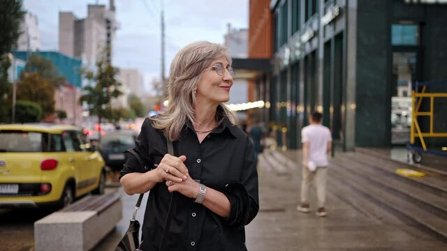 Happy mature woman with glasses and gray hair walks smiling along the city street. Walking from work or on a date
