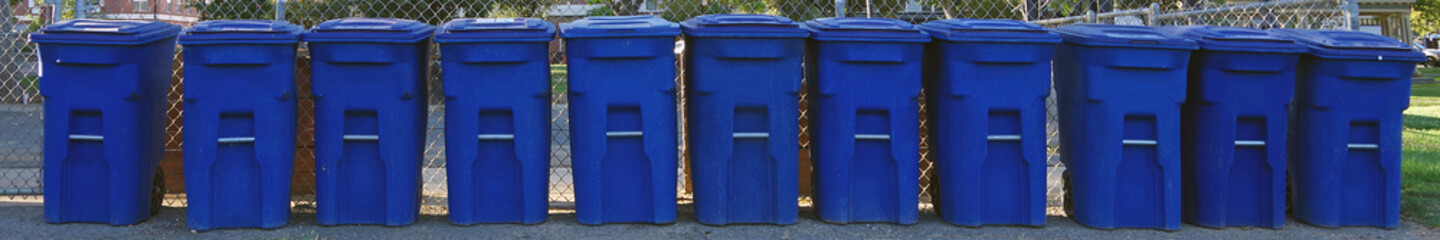 A long row of blue recycle trash containers lined up side by side along a chainlink fence