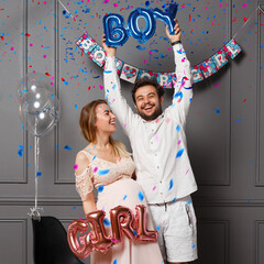 Happy couple holding balloons with inscription boy or girl during gender reveals party, over confetti and balloons.