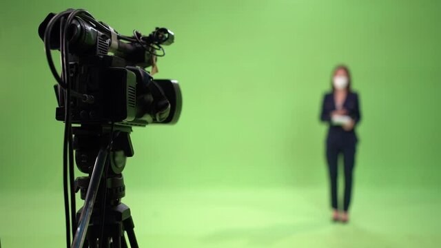 Selective focus of a video camera on a tripod are recording a blurred Asian female reporter in suit and wearing a face mask to protect Covid-19 standing talking on a green screen background in studio.