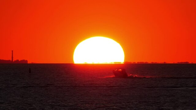 Large sun disk partially behind horizon, small motorboat race across bay and then stop, high contrast shot. Scenic telephoto view of sunset at Neva Bay in end of summer time
