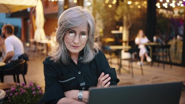 Gray-haired attractive business woman, a business owner, works in the evening in a city cafe. Mature lady carefully looks at the monitor of her laptop