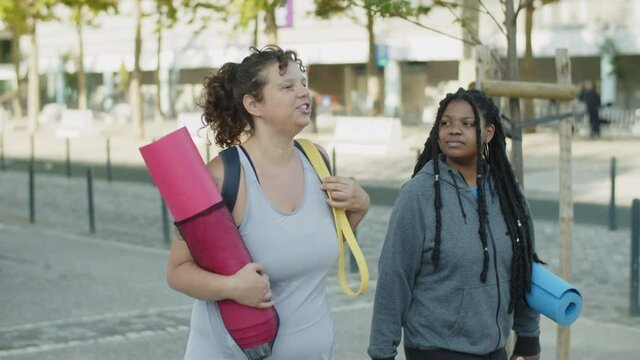 Diverse friends walking home through city streets after training. Tracking shot of fat friends with yoga mats in sportswear talking, spending weekend together. Body positive, friendship, sport concept