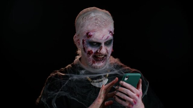 Frightening man with Halloween zombie bloody wounded makeup using mobile phone typing new post on web, sms message, browsing, addiction of social networks. Sinister undead guy on black wall background