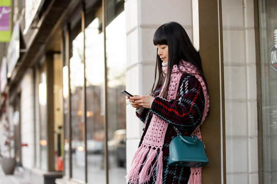 Stylish woman in pink scarf using smart phone at urban storefront