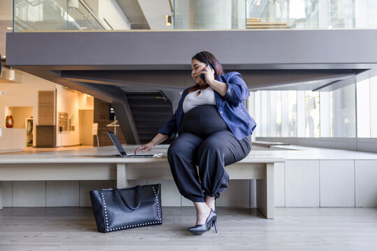 Businesswoman talking on smart phone at laptop in modern office lobby