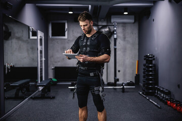 Fototapeta na wymiar Setting parameters of EMS training on a tablet. Young attractive man holds a tablet in the gym with a special EMS tablet while standing at the gym in an EMS suit. Electrical muscle stimulation