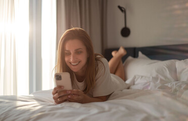 Happy woman lying on bed and using smartphone in bedroom.