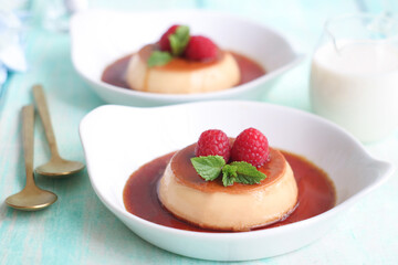 Plates of caramel custard pudding on table with two spoons, decorated with raspberries and mint leaf.