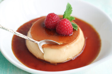 Close up of cream caramel custard  in a white plate, in a brown caramel sauce with spoon grabbing in a plate about to eat, with mint leaves  