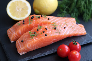 Fresh raw salmon with ingredient in preparation to cook with vegetable such as lemon, cherry tomatoes, peppers and dill on a black plate.Salmon having a lot nutrition benefits and good for health 