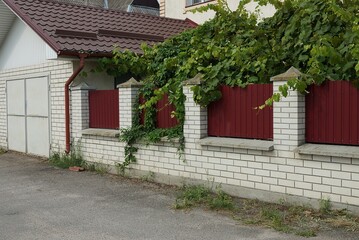 garage facade with gray iron gates against the wall of a fence made of white bricks and red metal on the street in green vegetation