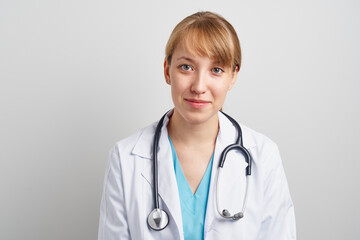 Young trustworthy woman healthcare worker with stethoscope and in white coat looking at camera and smiling. Portrait of confident family physician therapist on gray background.