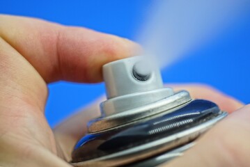 hand holds a black deodorant and a finger presses the gray button with a white spray on a blue background