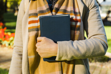 A girl in a warm scarf and a light coat clutches a blue book to her chest. Park at sunny day.
