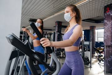 Two young beautiful girls exercise in the gym wearing masks during the pandemic. Social distancing in public places.