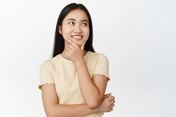 Obraz premium Smiling asian girl thinks, looks aside thoughtful, ponder something, standing over white background