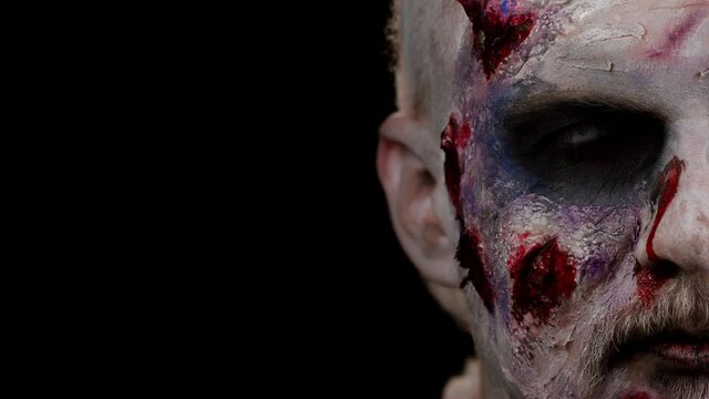Close-up zombie man face makeup with wounds scars and white contact lenses looking at camera, trying to scare in dark studio room background. Sinister dead guy. Halloween, filming, staging concept