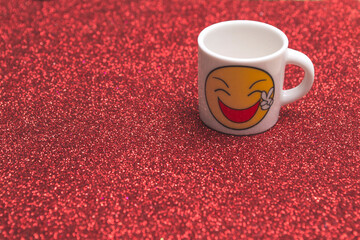 White cup with a smiling emoticon on a red glittering background. Day of smiles. copy space.