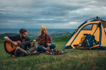 Couple of campers sitting near the tent while camping and playing guitar