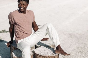 Fashionable african american man posing looking at camera in fashionable clothes outdoors while sitting on sand outdoors