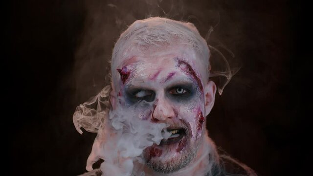 Sinister man with horrible scary Halloween zombie makeup in costume looking ominous at camera blows smoke from nose and mouth. Dead guy with wounded bloody scars face isolated against black background