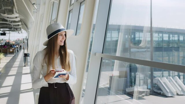 Elegant beautiful business girl holding travel tickets and using phone, happy girl looking at camera in airport terminal. Fashion woman portrait. Airport terminal. Business trip