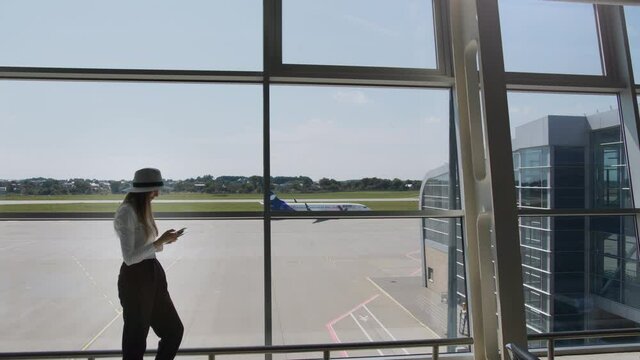 Young woman traveler, leisure or business standing at the glass window of a large international airport hub with a phone in his hands, scrolling the application for smartphones. Background of plane