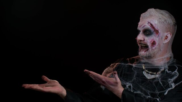 Creepy man with bloody scars face, Halloween stylish zombie make-up. Scary wounded undead guy showing thumbs up and pointing at left on blank space. Place for your advertisement logo. Sinister monster