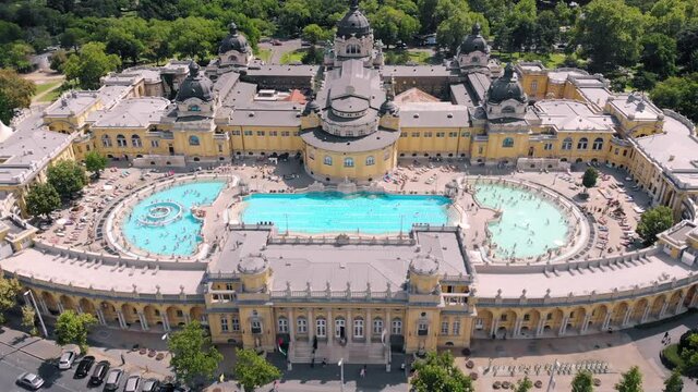 Szechenyi Bath aerial view. Drone flies above pool with many visitors. Camera moves back.