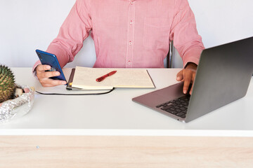young man at his office desk with his laptop in front of him, his mobile phone in his hand, a notebook in front of him with a red pen, on a white background, unrecognizable model