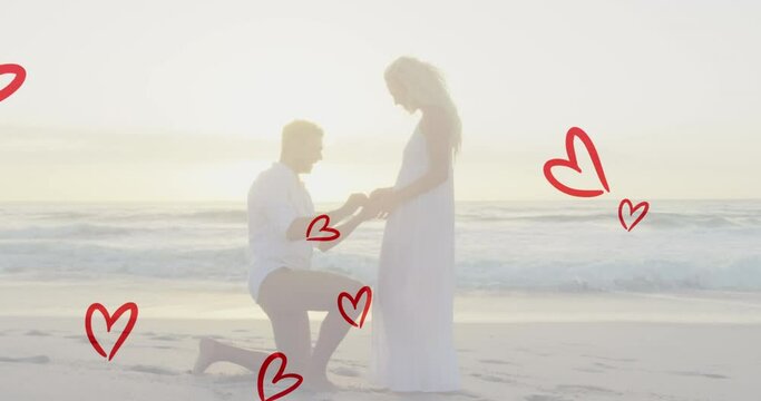 Animation of red hearts over caucasian man proposing on beach