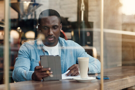 Purposeful black male entrepreneur uses tablet pc while sitting in cafe with coffee near window.