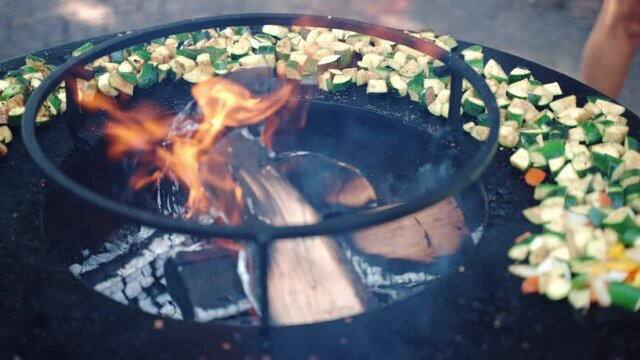 Black professional BBQ smoker with burning wood trunks and rising flames, cut zucchini, onion and bell pepper grilling in fire for weekend bbq picnic party. Process of cooking vegetables on grill grid