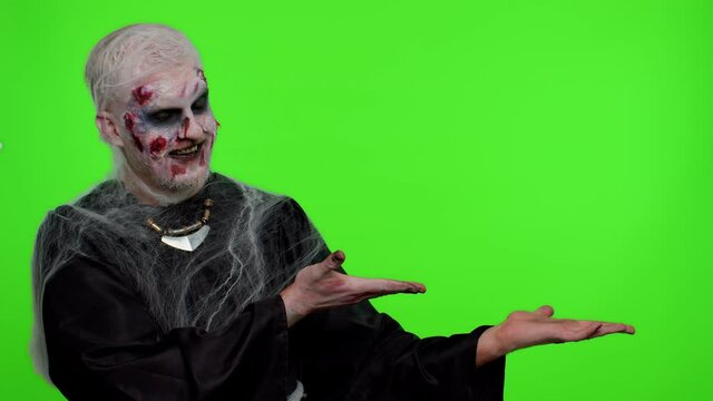 Creepy man with bloody scars face, Halloween stylish zombie make-up. Scary wounded undead guy showing thumbs up and pointing at right on blank space. Place for advertisement logo. Sinister monster