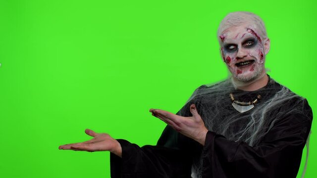 Creepy man with bloody scars face, Halloween stylish zombie make-up. Scary wounded undead guy showing thumbs up and pointing at left on blank space. Place for your advertisement logo. Sinister monster