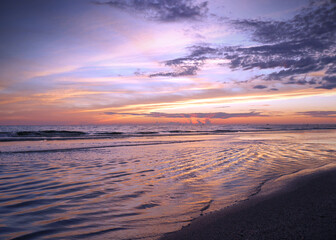 A post sunset sky of orange pink and purple hover over a calm shore with colors reelecting off of pools fo sea water collecting in the sand.