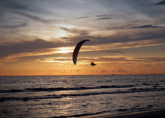 A silhouette of a person flying an ultralight in front of an sunset over the Gulf of Mexico.  The sky filed with flying seagulls and clouting clouds reflecting yellow, orange, and blue.