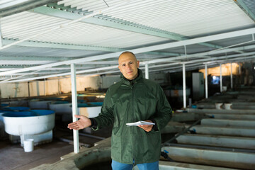 Confident farmer engaged in industrial breeding of trout, demonstrating fry rearing tanks on his farm..