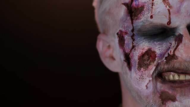 Close-up zombie man face makeup with wounds scars and white contact lenses, blood flows and drips on face trying to scare, face expressions in dark studio room background. Sinister dead guy. Halloween
