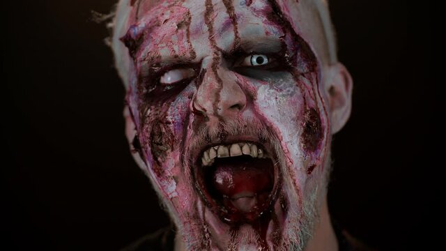 Close-up spooky zombie man face makeup with wounds scars, blood flows and drips on face trying to scare, screaming, shouting from pain in black studio room background. Sinister dead guy. Halloween