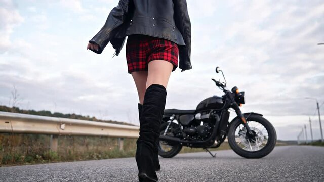 Unrecognizable motorcyclist woman in mini skirt and leather jacket walking to retro-styled motorcycle. Sexy female driver in jackboots alone on roadway. Trip, cafe racers, speed, freedom concept.