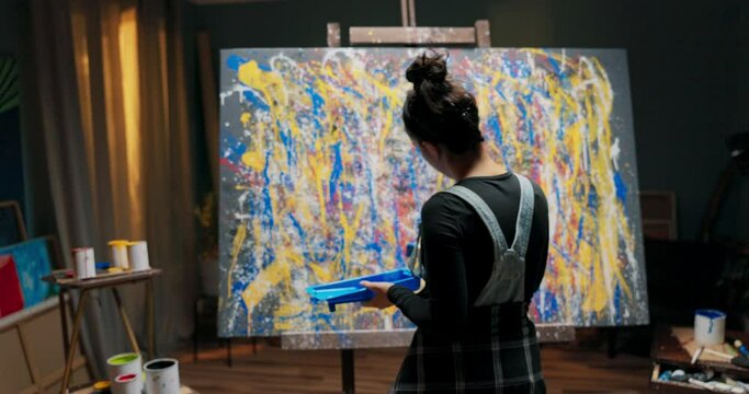 Dark studio lit by lamp at night woman artist painter stands by canvas with abstract painting in hand she holds container with blue paint and brush applies with quick movement