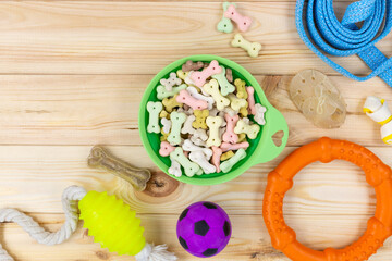 Different multicolored pet care accessories: green bowl, bones, balls, snacks on natural wooden background. Rubber and textile accessories for dogs. Top view, flat lay. Copy space	