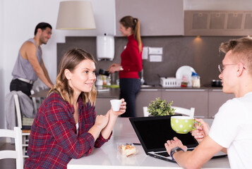 Attractive girl sitting in kitchen of hostel with cup of coffee, talking to guy with laptop