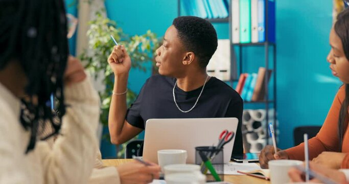 Young handsome man with dark skin sits in middle of coworkers, discusses vision for project, looks at laptop, talks, shows a sample graphic of the logo to everyone right and left on the tablet