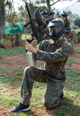 Fototapeta premium Young woman in camouflage holding gun before paintball match with friends on open field