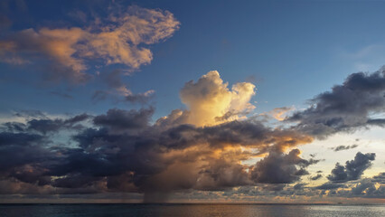 There are dark blue and golden clouds in the azure evening sky. In the distance, over the ocean, it's raining. Reflection on the surface. Maldives