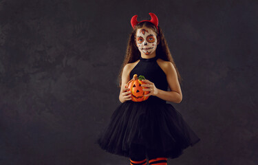 Kid in spooky costume on dark background. Studio shot of young child on Halloween. Girl wearing beautiful black dress, red devil horns and skull makeup holding orange pumpkin and looking at camera
